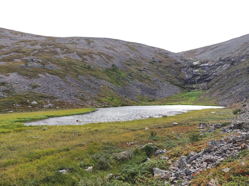 Paratiisikurun pohjalla oleva lampi, jonka ranta on vihreää ruohikkoa. Takana näkyy kiviset tunturinrinteet ja vesiputous.